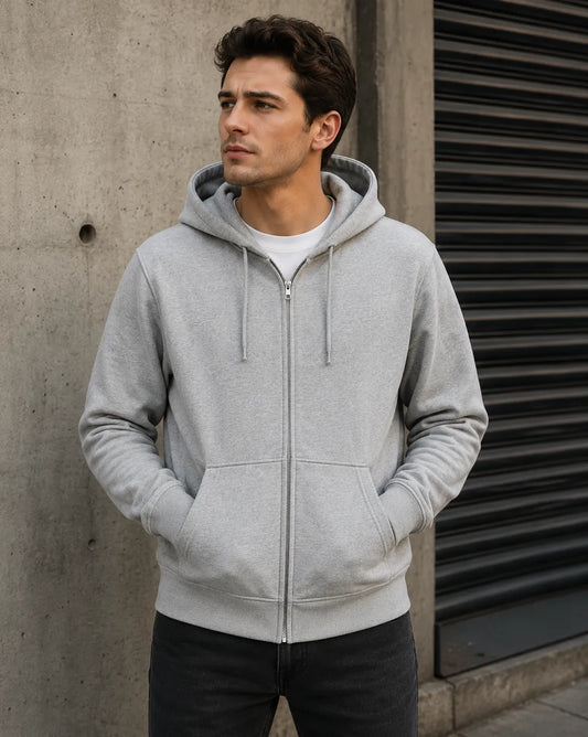 Man wearing a gray hoodie standing against a concrete wall.